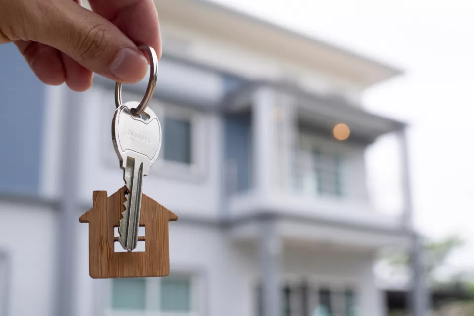 hand holding house key with house-shaped keychain and house in background